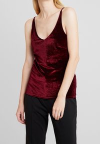 Camisola di velluto bordeaux con scollatura a V profonda, spalline sottili e una consistenza morbida e lussuosa. Abbinata a pantaloni neri larghi.
