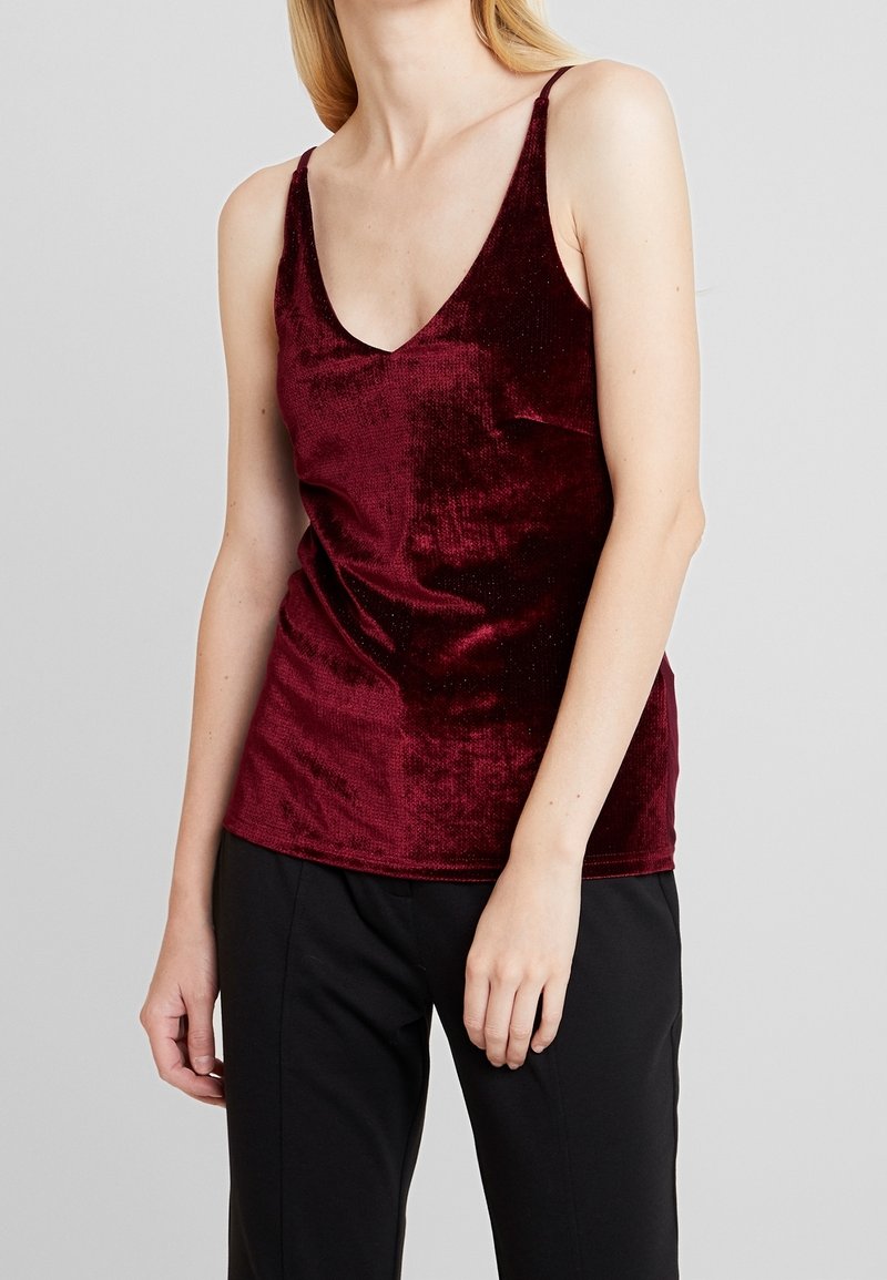 Camisola di velluto bordeaux con scollatura a V profonda, spalline sottili e una consistenza morbida e lussuosa. Abbinata a pantaloni neri larghi.
