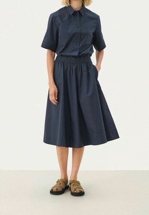 Osoba ubrana w granatową koszulę na krótki rękaw z guzikami, dopasowaną midi spódnicę z elastycznym pasem oraz brązowe sandały z odkrytymi palcami, stojąca na białym tle.