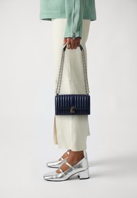 Navy quilted leather handbag with a chain strap, rectangular shape, featuring a silver-tone clasp. Model wears cream pants and silver shoes.