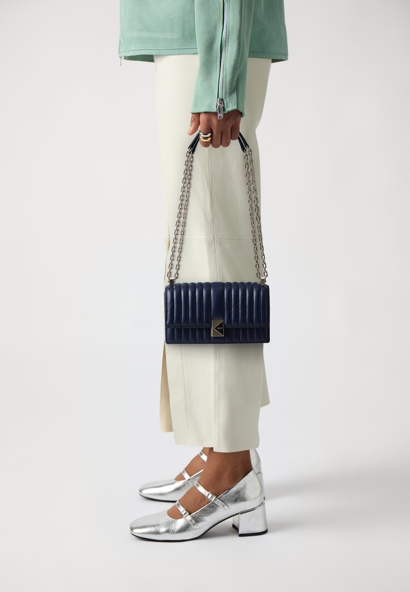 Sac en cuir matelassé bleu marine avec une bandoulière en chaîne, de forme rectangulaire, doté d'un fermoir en métal argenté. Le modèle porte un pantalon crème et des chaussures argentées.