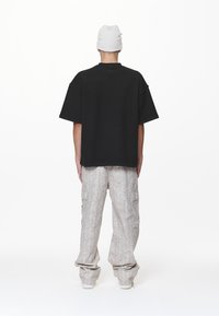 Persoon in een witte beanie, losse zwarte shirt met korte mouwen en lichtgrijze cargo broek met patroon, staande met de rug naar de camera op een witte achtergrond.