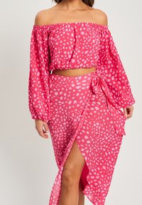 Růžový cropped top s odkrytými rameny a dlouhými rukávy, který je spárován s odpovídajícími zavinovacími sukněmi s bočním rozparkem a detailem na zavazování, obě s puntíkovaným vzorem.