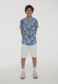 Camicia blu a maniche corte con stampa di palme bianche, abbinata a pantaloni corti bianchi strappati, calze bianche con strisce nere e scarpe chiare.