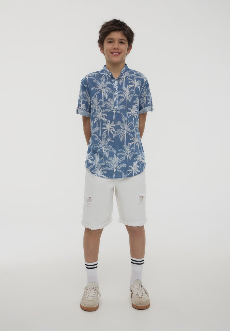 Camicia blu a maniche corte con stampa di palme bianche, abbinata a pantaloni corti bianchi strappati, calze bianche con strisce nere e scarpe chiare.