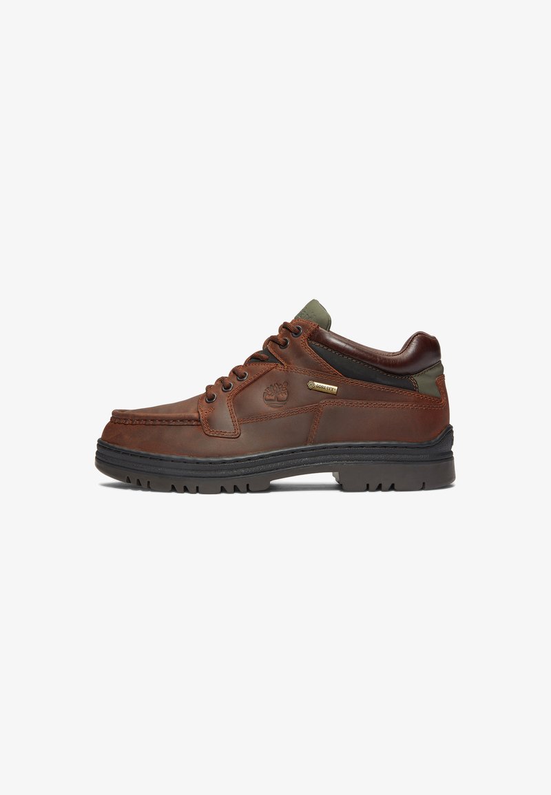 Zapato de cuero marrón con un diseño clásico, que presenta un cierre con cordones, detalles en verde y una suela de caucho negro resistente. Etiqueta GORE-TEX® visible.