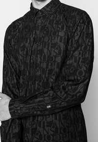 Camicia nera con bottoni realizzata in tessuto testurizzato con un motivo di testo audace ripetuto. Progettata con colletto classico e polsini aderenti.