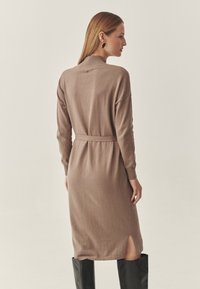 Neulottu pitkähihainen mekko taupen sävyssä, jossa on poolokaulus, vyötärövyö, sivuhalkiot ja resorit hihansuissa, yhdistetty polveen ulottuviin mustiin saappaisiin.