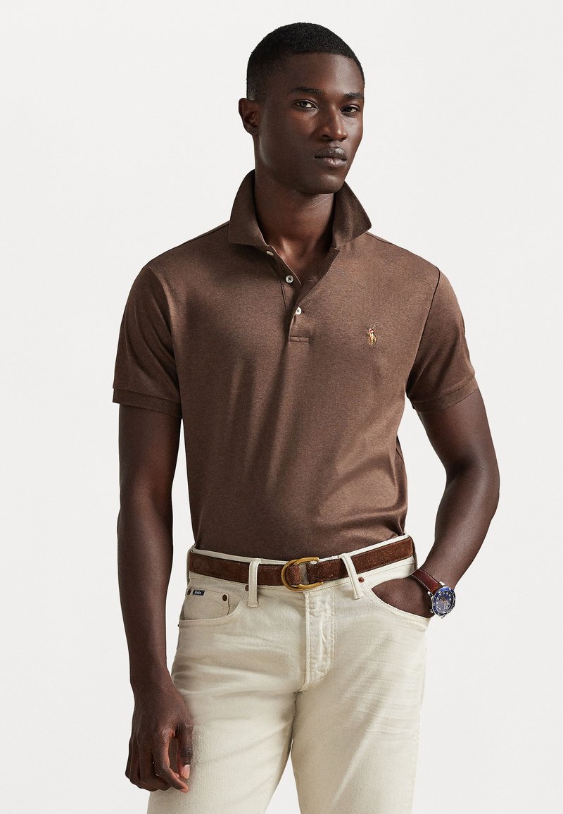 Camisa polo castanha de mangas curtas e colarinho dobrado, com um pequeno logótipo no peito. Usada com jeans de cor clara e um cinto castanho.