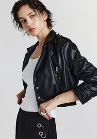 Schwarze kurze Lederjacke mit einer Reißverschlusstasche, Druckknöpfen und geripptem Saum. Getragen über einem weißen Tanktop mit einer glatten Textur.