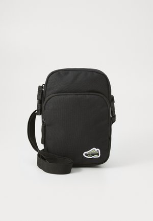 Bolso negro cruzado hecho de un tejido duradero, con un compartimento principal, correa ajustable y un detalle de parche en forma de zapatilla verde.