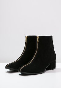 Bottines en daim noir avec fermetures éclair dorées à l'avant et talons blocs bas sur une surface blanche et un fond gris clair.