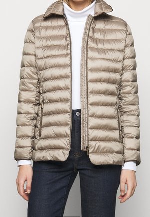 Mujer vistiendo una chaqueta acolchada marrón claro sobre un suéter de cuello alto blanco y jeans azul oscuro, con la chaqueta parcialmente desabrochada mostrando la cremallera y los bolsillos.