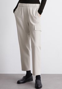 Hellbeige, taillierte Hose mit einem elastischen Bund, seitlichen Taschen und einer Cargotasche am linken Bein. Der Stoff wirkt glatt und dehnbar.