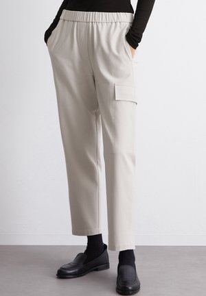 Pantalon tapered beige clair avec une taille élastique, poches latérales et une poche cargo sur la jambe gauche. Le tissu semble lisse et extensible.