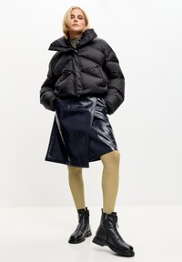 Giacca a vento nera, gonna avvolgente in finta pelle lucida blu navy, collant a coste colore senape, stivaletti neri con zip e caviglia, trame lisce, design corto.