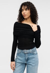 Μαύρο τοπ έξω από το ώμο με υφασμένο πλεκτό μοτίβο, σε συνδυασμό με τζιν παντελόνι υψηλής μέσης σε ανοιχτό μπλε και ζώνη.