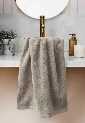 Toalla gris colgada sobre una encimera de lavabo de mármol blanco con grifo dorado, espejo redondo, planta en maceta, dispensador de jabón y vela encendida.
