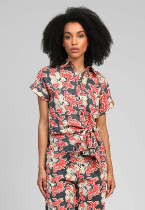 Femme aux cheveux bouclés naturels portant un ensemble chemisier à manches courtes et pantalon assortis à imprimé floral rouge, crème et noir, avec une ceinture à la taille.