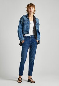 Chaqueta de mezclilla en azul claro, camiseta blanca con logo, jeans azul oscuro y sandalias de tacón negras con tiras. Look casual con un estilo de capas.