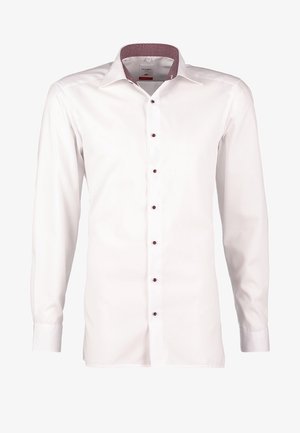 Homme marchant portant une veste en cuir noire, une chemise blanche boutonnée, un pantalon beige et des baskets blanches sur un fond uni.