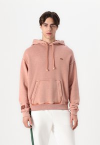 Sudadera con capucha en color rosa empolvado, hecha de suave tela de algodón. Cuenta con un bolsillo frontal, cordón ajustable y un pequeño logo en el pecho.