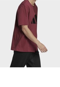 Bordeaux t-shirt i bomuld med korte ærmer, prydet med et stort sort logo foran. Modellen har sorte bukser på og er fanget i en gående positur.