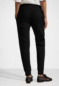 Černé teplákové kalhoty s úzkým střihem, elastickým páskem, bočními kapsami a jednou zadní kapsou. Textura působí měkce a pohodlně.