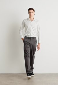Vit skjorta med knappar, gråbyxor, svarta sneakers. Slim fit, mjuk textur, definierade sömmar och minimalistisk design. Enfärgad bakgrund.
