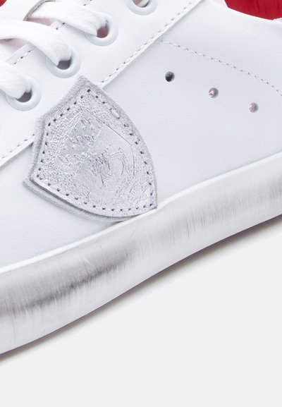 Λευκό δερμάτινο αθλητικό παπούτσι με γκρι λογότυπο σε σχήμα ασπίδας με υφή, σχέδιο με κορδόνια και σόλα με ασημί λεπτομέρειες που φέρει μια διακριτική φθορά.