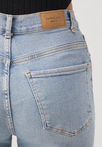 Světle modré džínové kalhoty, pohled zezadu, s viditelnou kapsou a hnědou koženou značkovou nášivkou s nápisem "perfect jeans gina tricot."