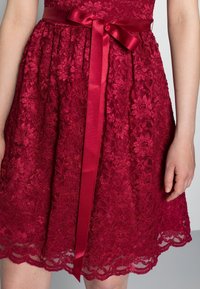 Robe en dentelle rouge ornée de motifs floraux, corsage ajusté, et ceinture en satin à la taille avec un ourlet festonné.