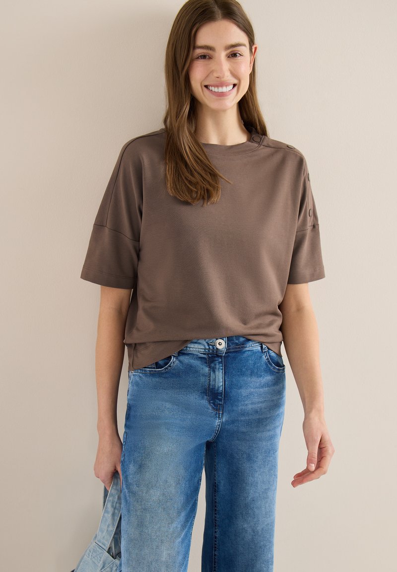 Jeune femme souriante, portant une chemise marron à manches courtes et un jean bleu, tenant un sac fourre-tout en denim assorti sur un fond neutre.
