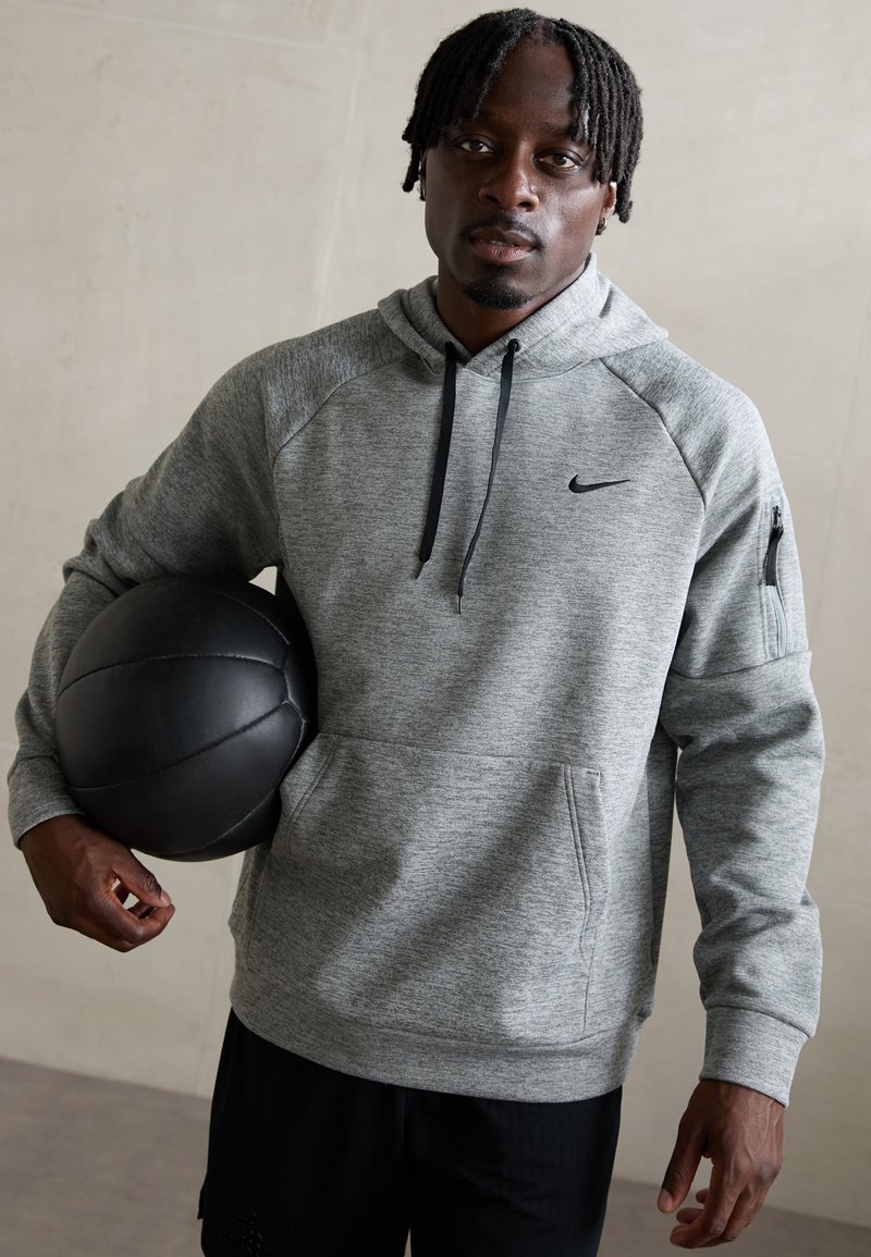 Grå meleret hoodie med en frontlomme, sidelomme med lynlås og sorte detaljer, båret mens der holdes en sort basketball.
