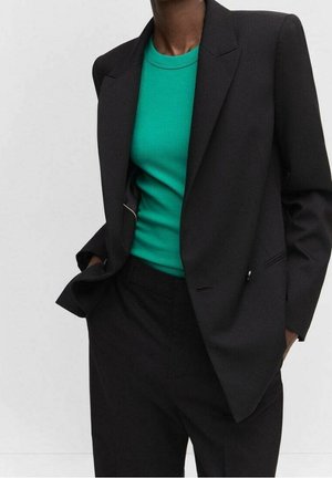 Personne portant un blazer noir et un pantalon avec un pull côtelé vert vif à col rond, posant les mains dans les poches devant un fond uni.