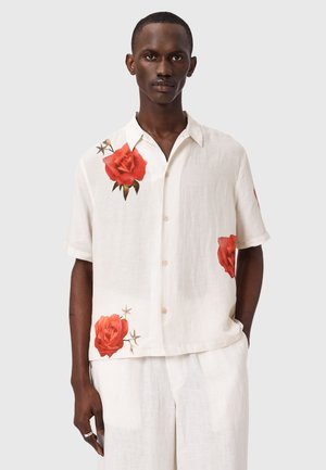 Mand iført hvid kortærmet skjorte med rød roseprint på skulderen, nederste forside og side, kombineret med hvide bukser.