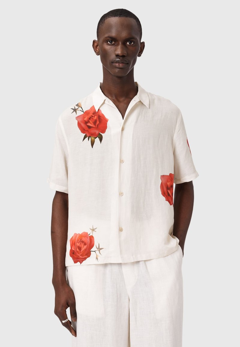 Hombre con camisa blanca de manga corta y botones, con estampados de rosas rojas en el hombro, parte inferior delantera y lateral, combinada con pantalones blancos.