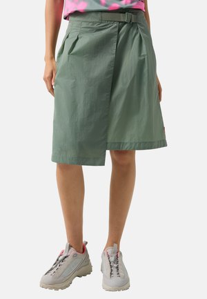 Jupe portefeuille verte longueur genou avec ceinture portée avec des baskets roses et grises, présentée sur une personne debout sur un fond uni.