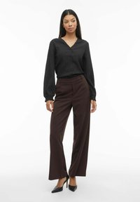 Melna blūze ar gariem piedurknēm, vertikālām svītrām un pogu aizdari priekšpusē, kopā ar augstajiem brūniem plašām biksēm un melniem smailiem papēžiem.