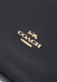 Czarna teksturowana skórzana powierzchnia z złotym logo Coach New York, zawierającym emblemat konia i powozu.