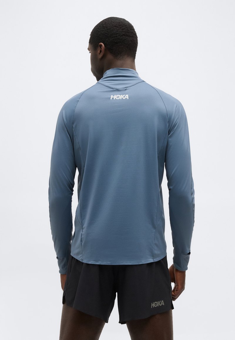 Modelo masculino vistiendo una camiseta deportiva azul de manga larga y pantalones cortos de correr negros, ambos con logotipos blancos de "HOKA", visto desde atrás.