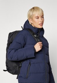 Jūras spēku pufīgā jaka ar quilted tekstūru, ar kapuci un ar rāvējslēdzējiem aprīkotām kabatām. Valkāta ar melnu mugursomu, kas aprīkota ar regulējamām siksnām.