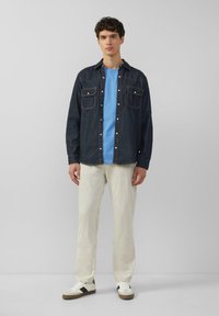 Jonge man die staat, gekleed in een donkerdenim overhemd met knopen over een blauwe t-shirt, crèmekleurige broek en witte sneakers met zwarte strepen.