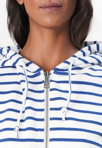 Sweat à capuche zippé blanc avec des rayures horizontales bleues ; il est doté d'une capuche à cordon et d'un accent en métal sur la fermeture éclair. Matière en mélange de coton doux.