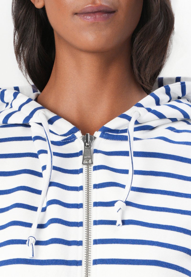 Hoodie z zamkiem błyskawicznym w białym kolorze z niebieskimi poziomymi paskami; posiada kaptur z regulacją sznurkiem oraz metalowy akcent w postaci zamka. Wykonany z miękkiej mieszanki bawełny.