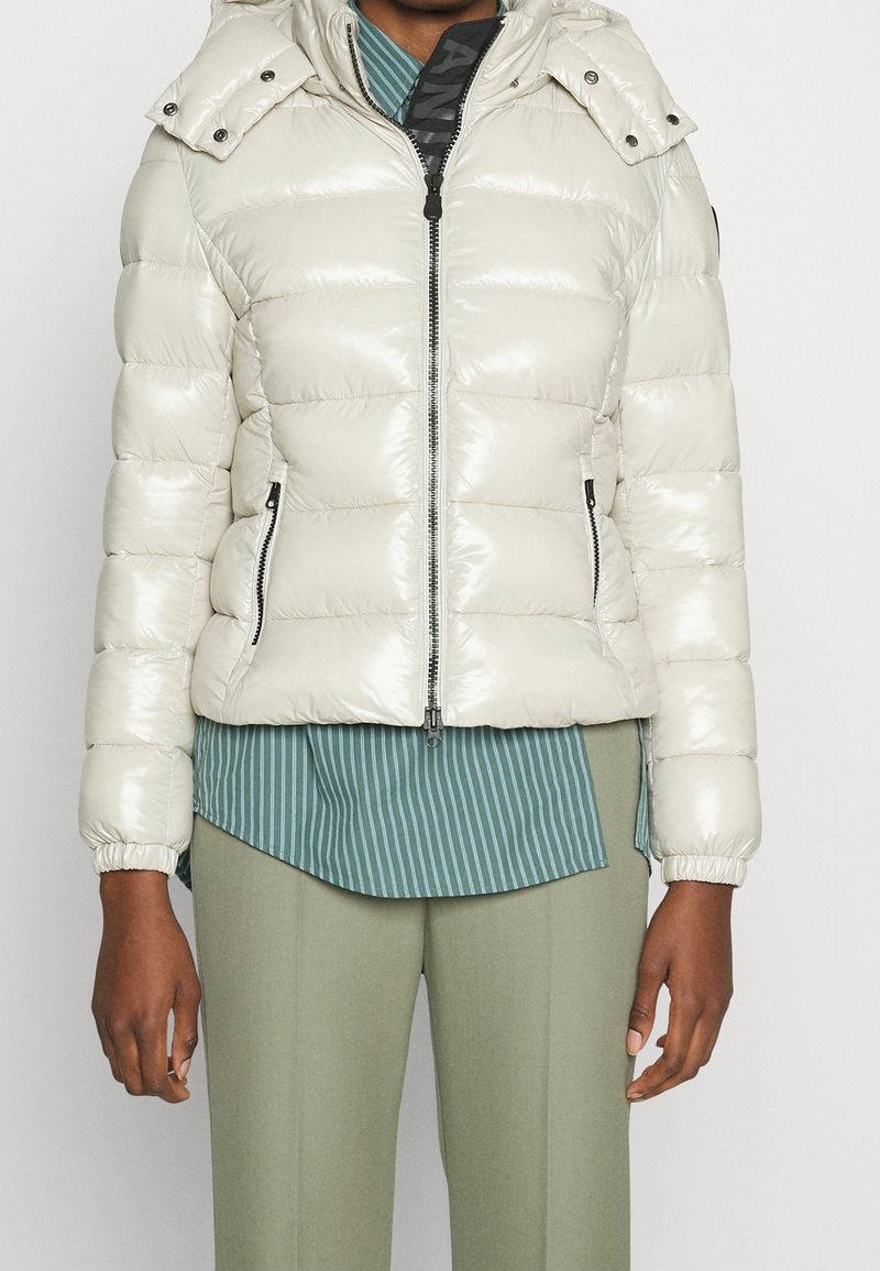 Personne portant une doudoune brillante beige clair avec des poches zippées, sur une chemise à rayures vertes et un pantalon vert clair.