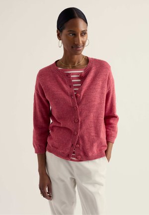 Vrouw draagt een knoopsgat roze-roze vest over een roze-wit gestreept shirt, met witte broek, gouden ketting en oorringen.