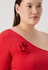 Rød tettsittende topp med asymmetrisk utringning og en teksturert roseaccent på skulderen. Glatt, elastisk materiale.