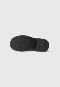Semelle de chaussure noire avec grip texturé, pièce de talon rectangulaire fixée par quatre rivets visibles, présentée sur un fond clair uni.