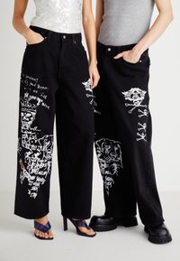 Jeans en denim noir à large jambe présentant des impressions graphiques blanches de crânes et de chiens, avec deux poches avant et une coupe décontractée.